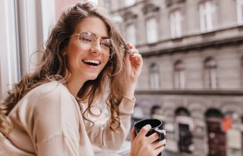 A woman with long hair and glasses smiles while holding a black mug, sitting by a window overlooking a street., 